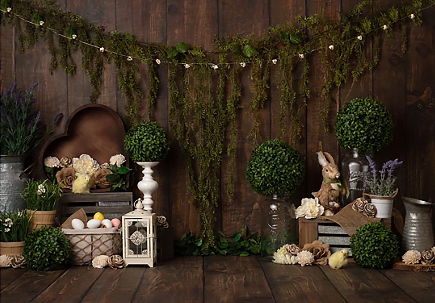 Easter backdrop with green topiaries, bunny figurines, eggs, lantern, and string lights on rustic wood wall.