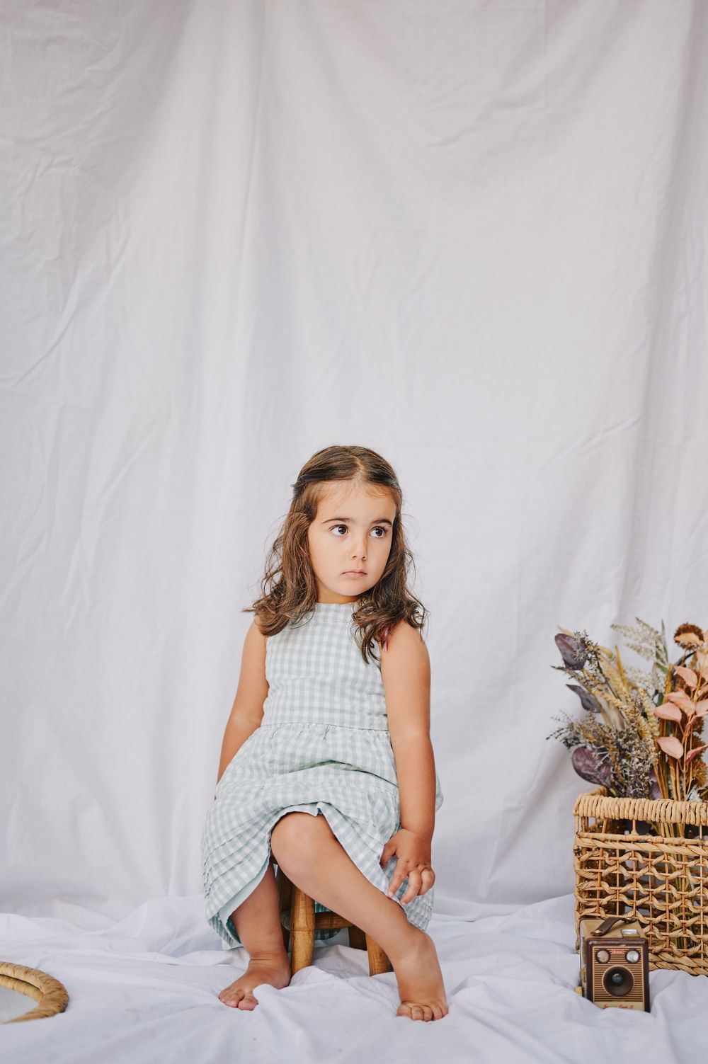 Young girl in sage gingham dress sitting on stool against white backdrop.