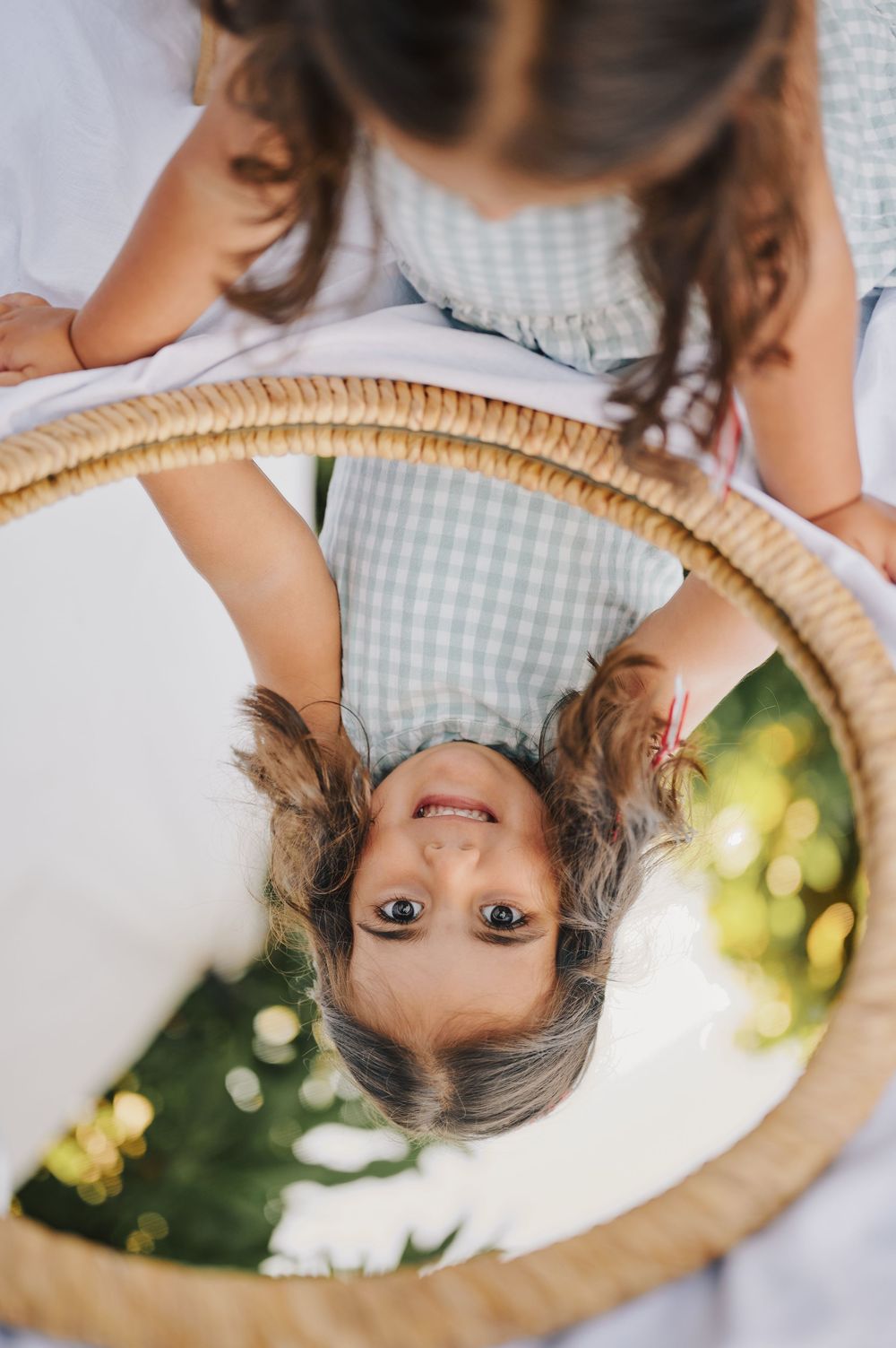 Child reflected upside down in round mirror held by older girl outdoors.