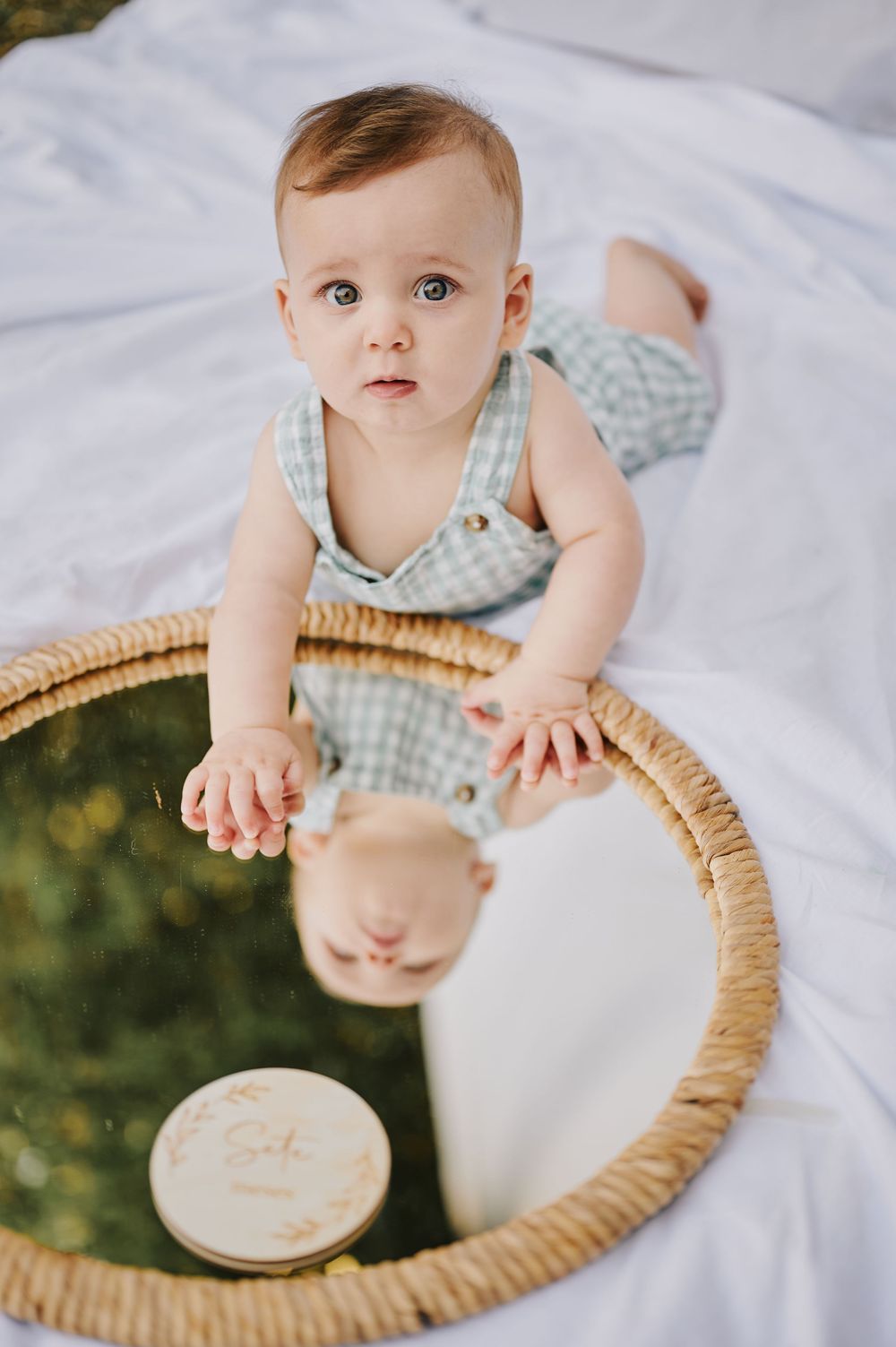 Curious baby in a green romper leaning over a round rattan mirror on a white blanket outdoors.