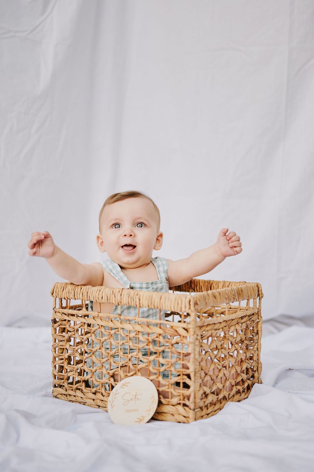 Happy baby sitting in a large wicker basket with arms raised, smiling against a white backdrop.