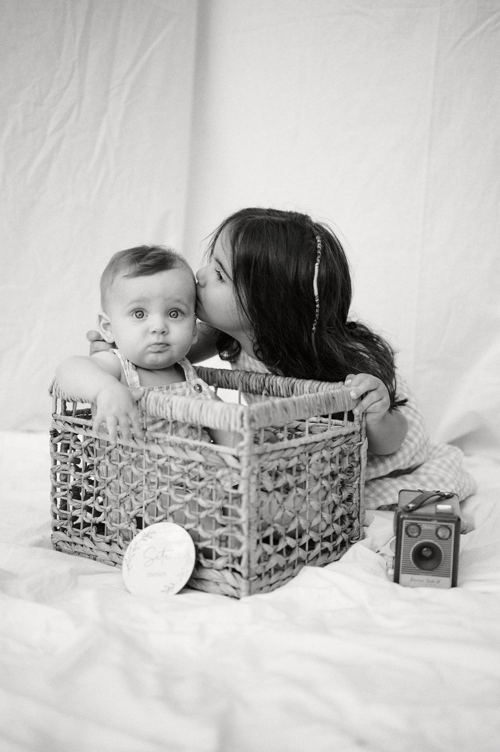 Black and white photo of older sister kissing baby sibling in wicker basket.