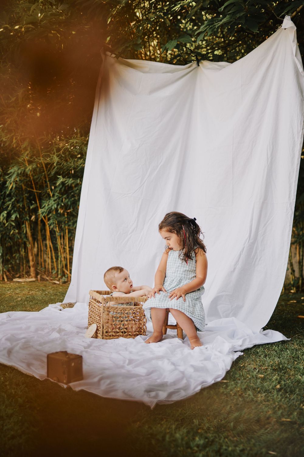 Young girl in gingham dress playing with baby in wicker basket outdoors.