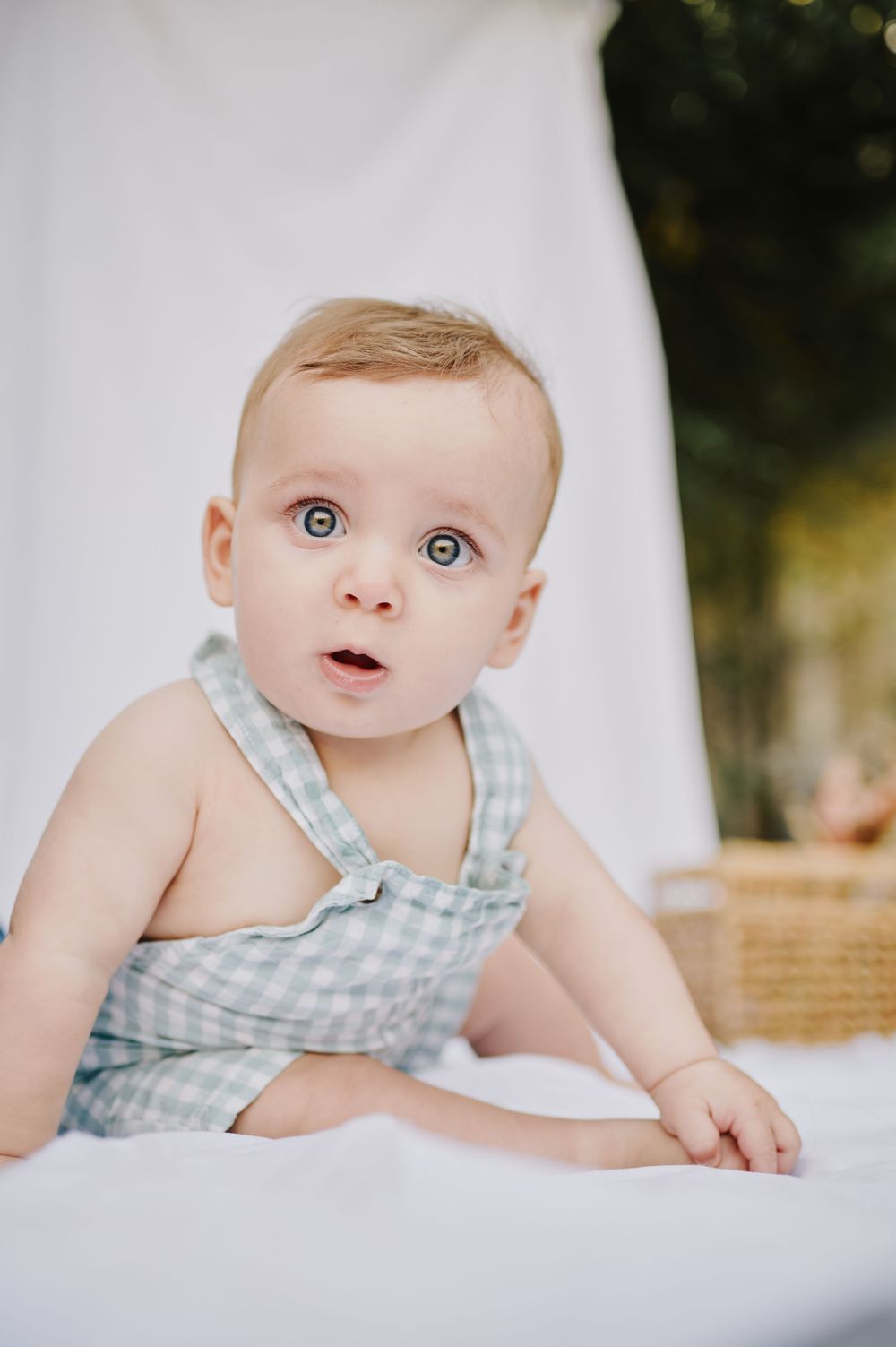 Curious baby in gingham overalls lying on white fabric outdoors.