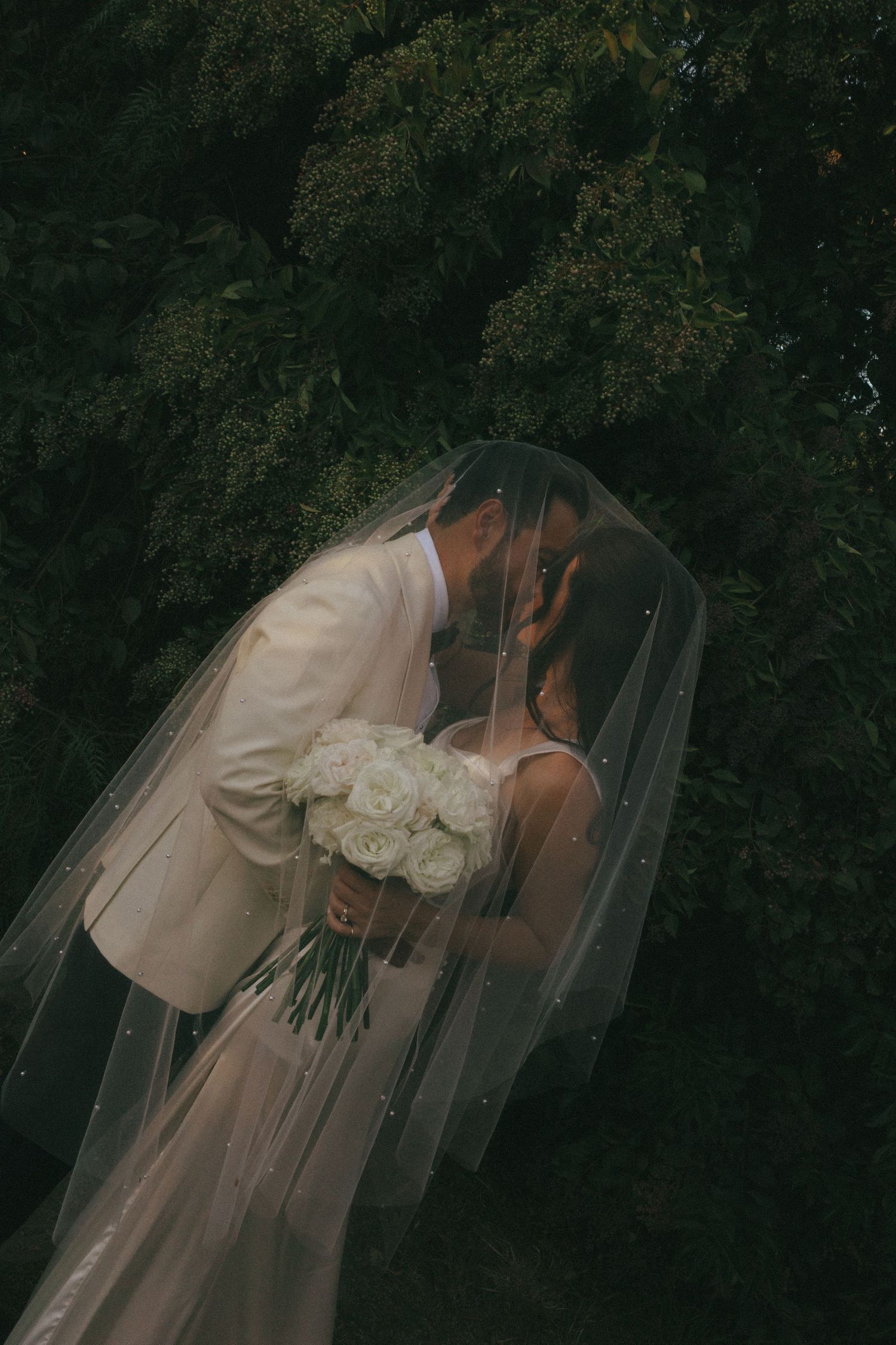 A romantic wedding moment captured in dark moody lighting as the couple shares a kiss under a flowing white veil.