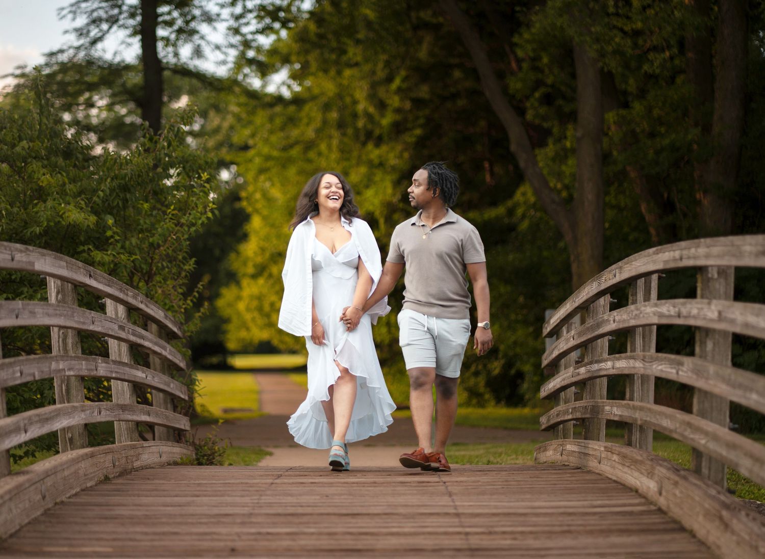 A couple walks hand in hand across a wooden footbridge in a lush green park setting.