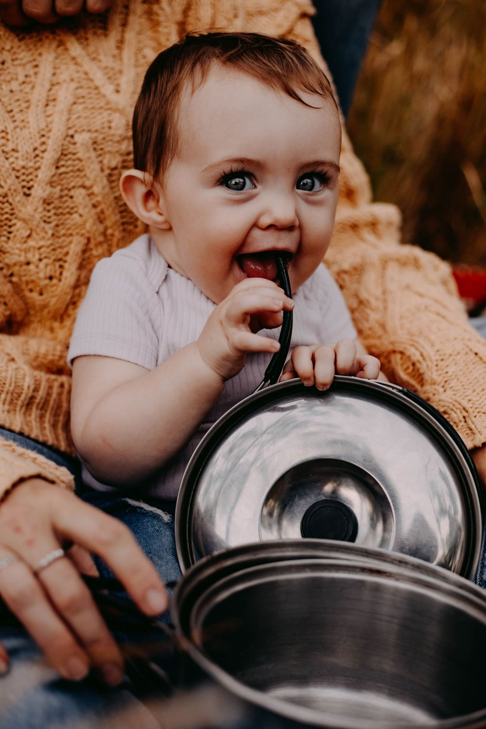 Baby with bright eyes plays with metal pots and pans while sitting on a knitted blanket.