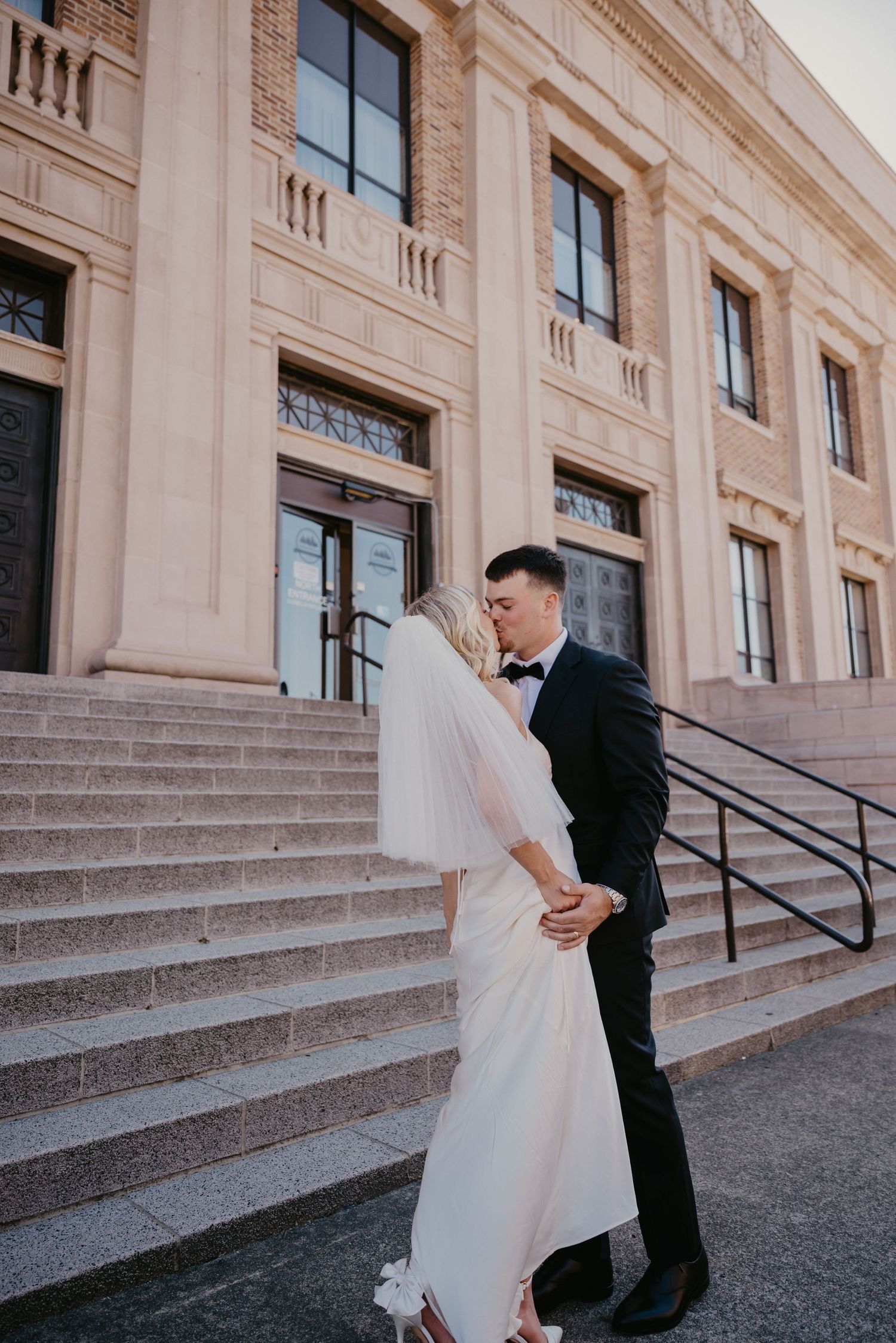 A bride and groom share a romantic moment on the grand steps of an elegant stone building.