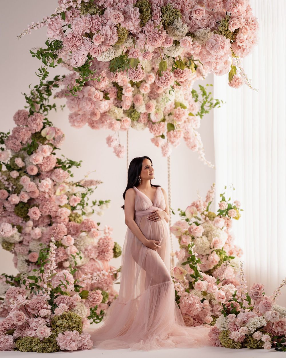 Elegant maternity photo featuring a flowing pink dress and cascading cherry blossom floral arch against a light backdrop.