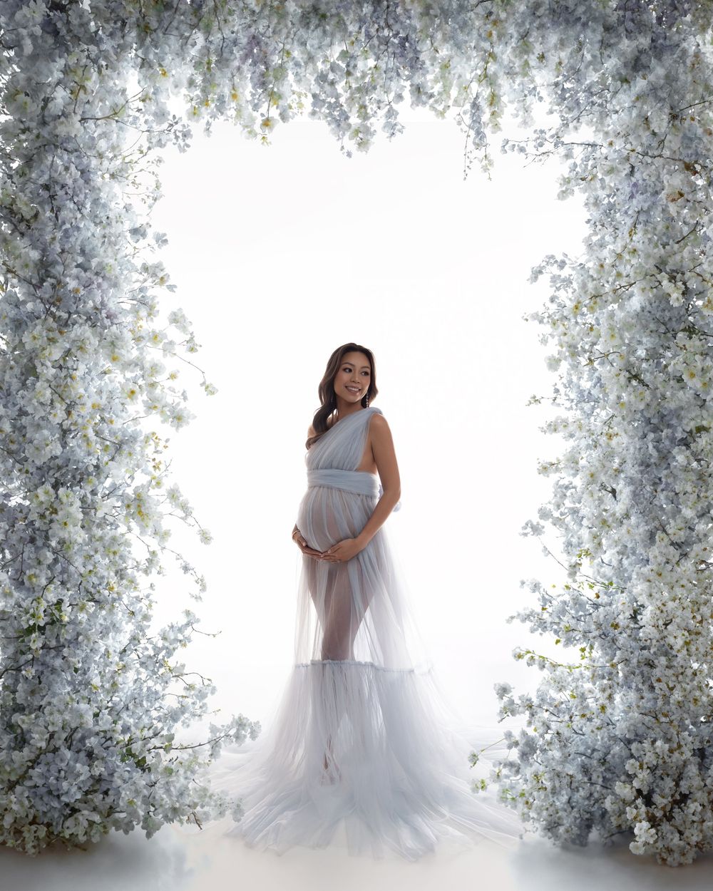 Pregnant woman in a flowing blue-gray tulle gown standing under a floral arch of white and blue blooms.