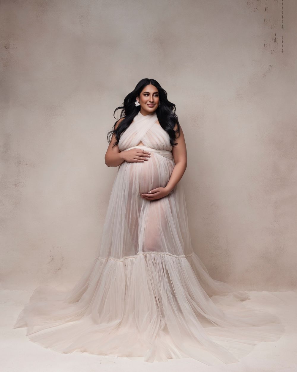 Pregnant woman in sheer white tulle maternity gown poses gracefully in a professional studio photo shoot.