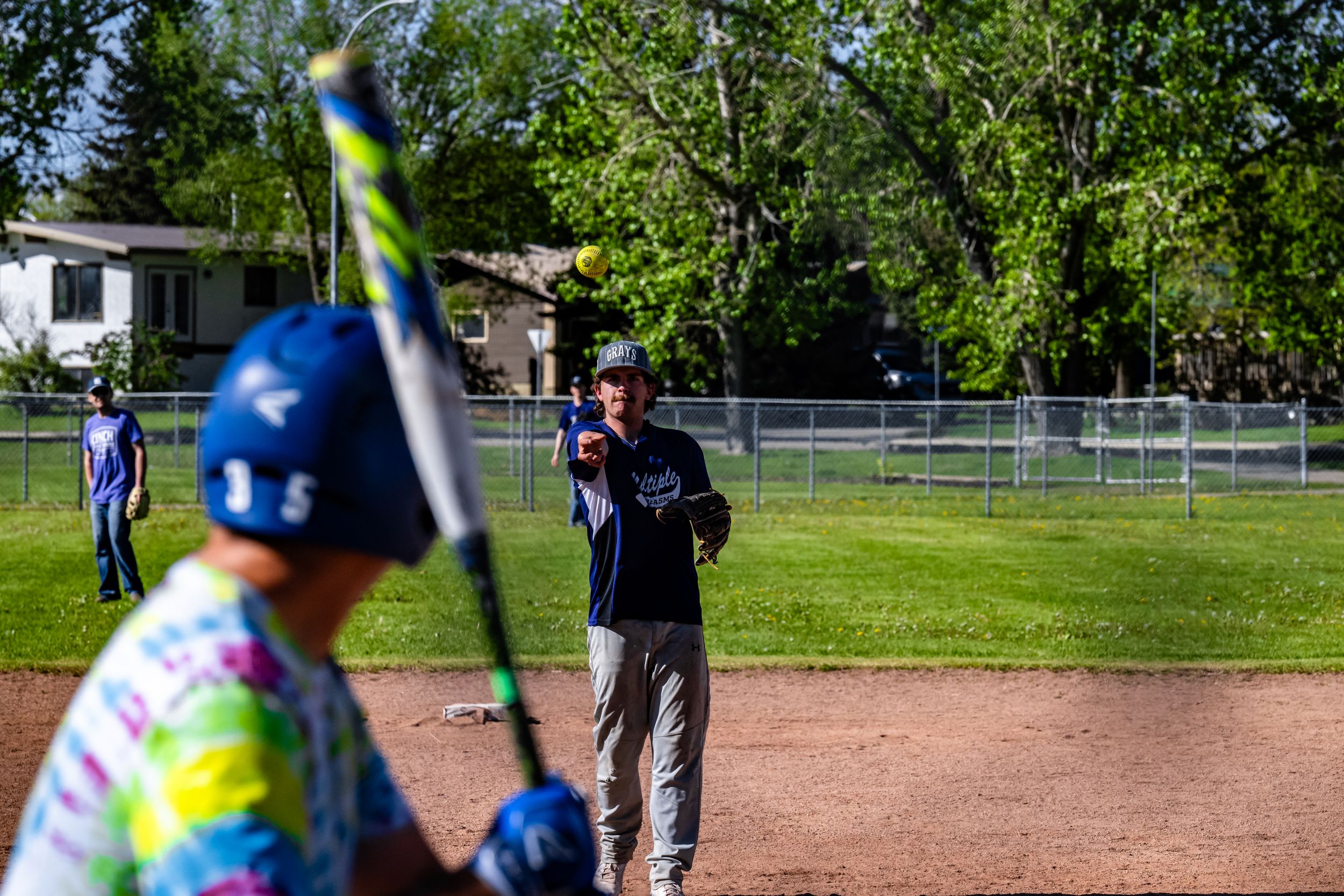 Baseball player in blue helmet and tie-dye jersey swings bat during game on sunny day at dirt baseball field.