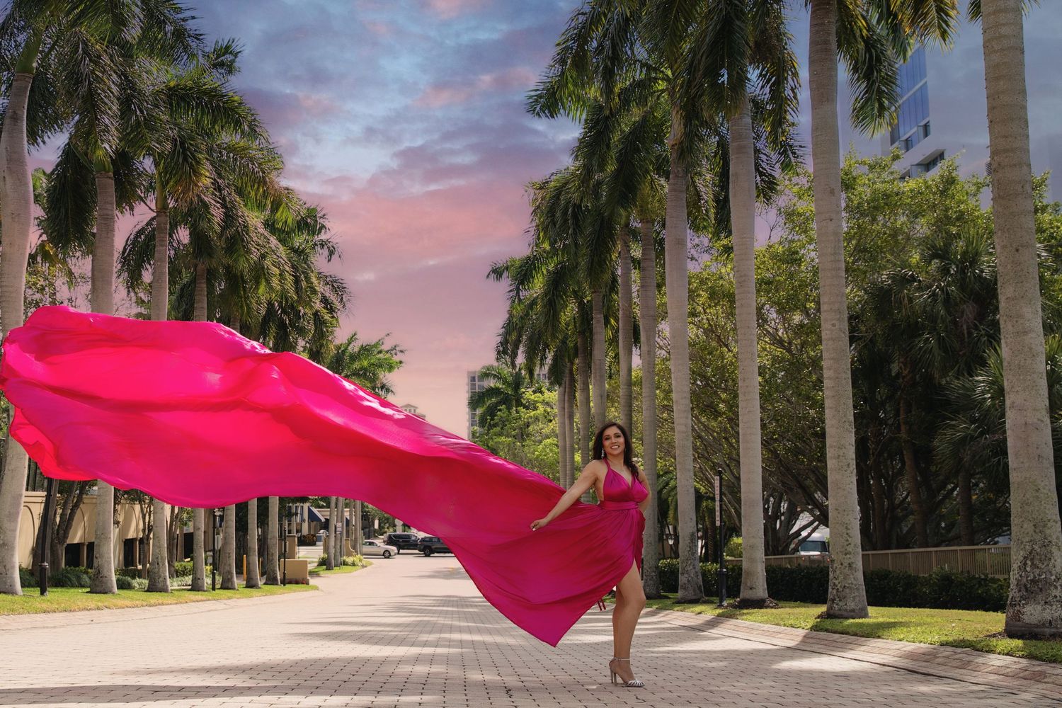 Pink kites soar between rows of tall palm trees lining a path during sunset with colorful clouds in the sky.