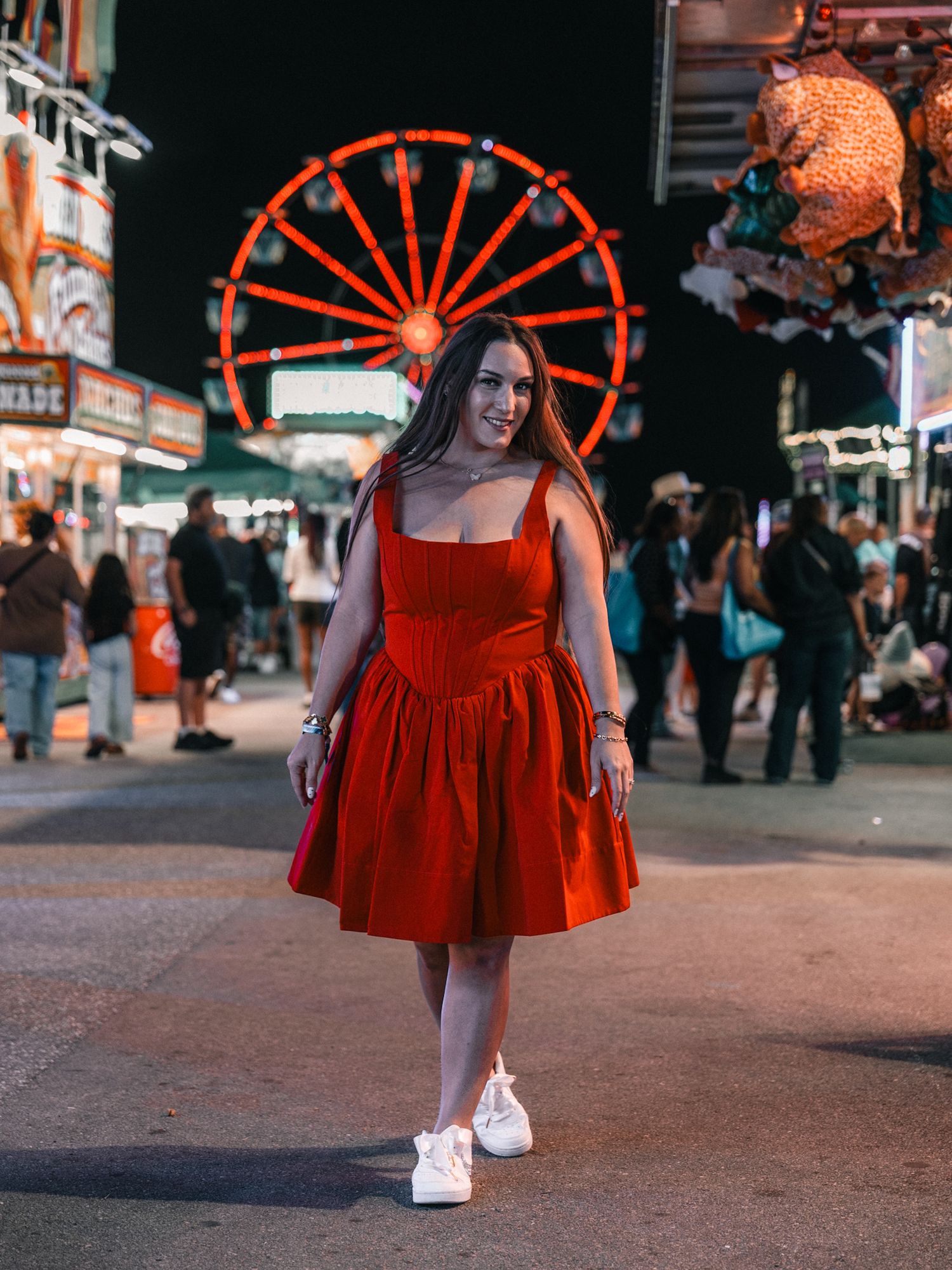 A person in a vibrant red dress poses at a carnival at night with a illuminated Ferris wheel in the background.