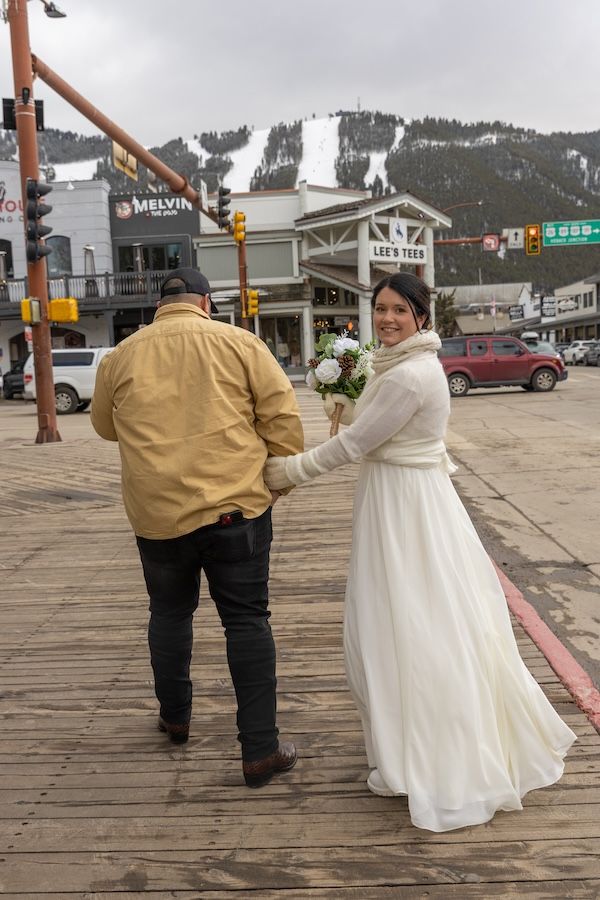 A couple in winter wedding attire walk along a wooden boardwalk with snowy ski slopes visible in the mountain background.