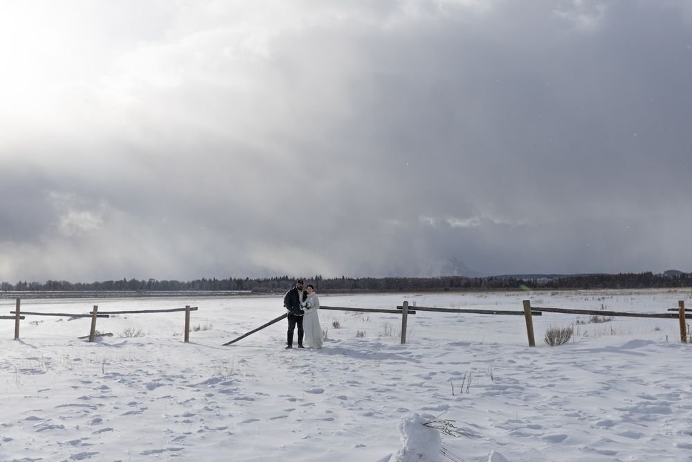 A person and dog stand in a snowy winter landscape with wooden fencing and stormy clouds overhead.