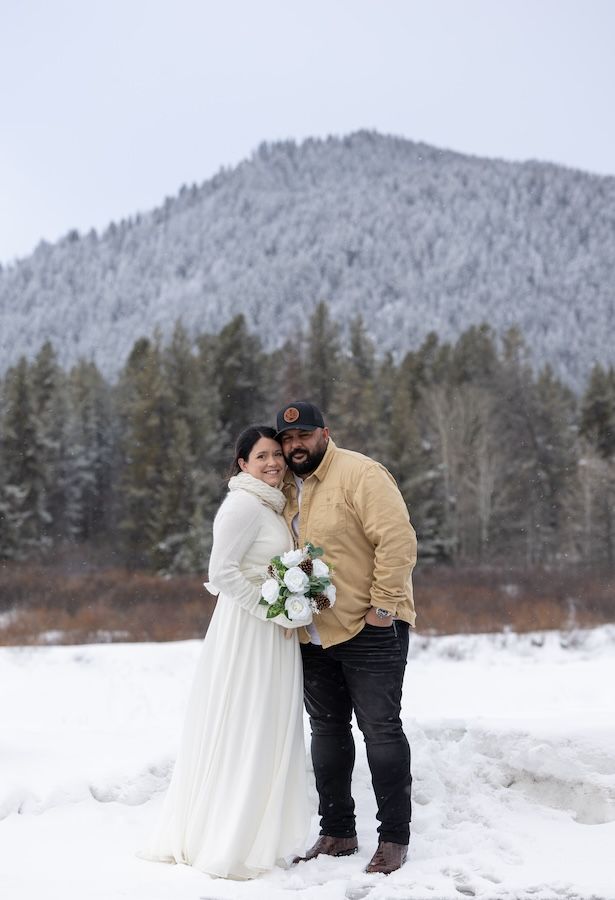 A couple shares an intimate winter moment in the snow with mountains and evergreen trees creating a romantic backdrop.