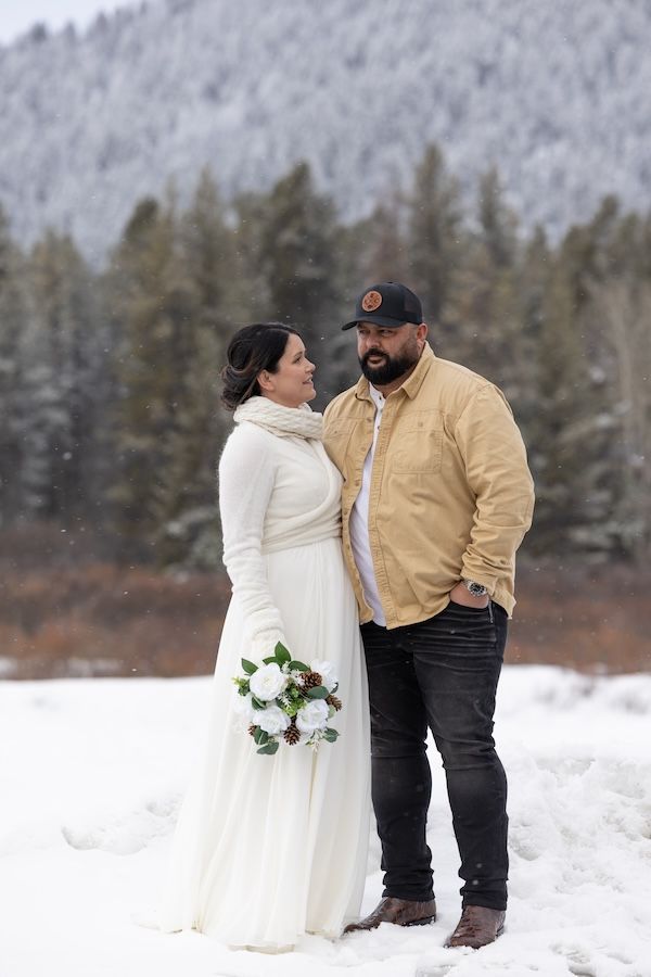 A couple shares a romantic moment in a snowy winter landscape with evergreen trees in the background.