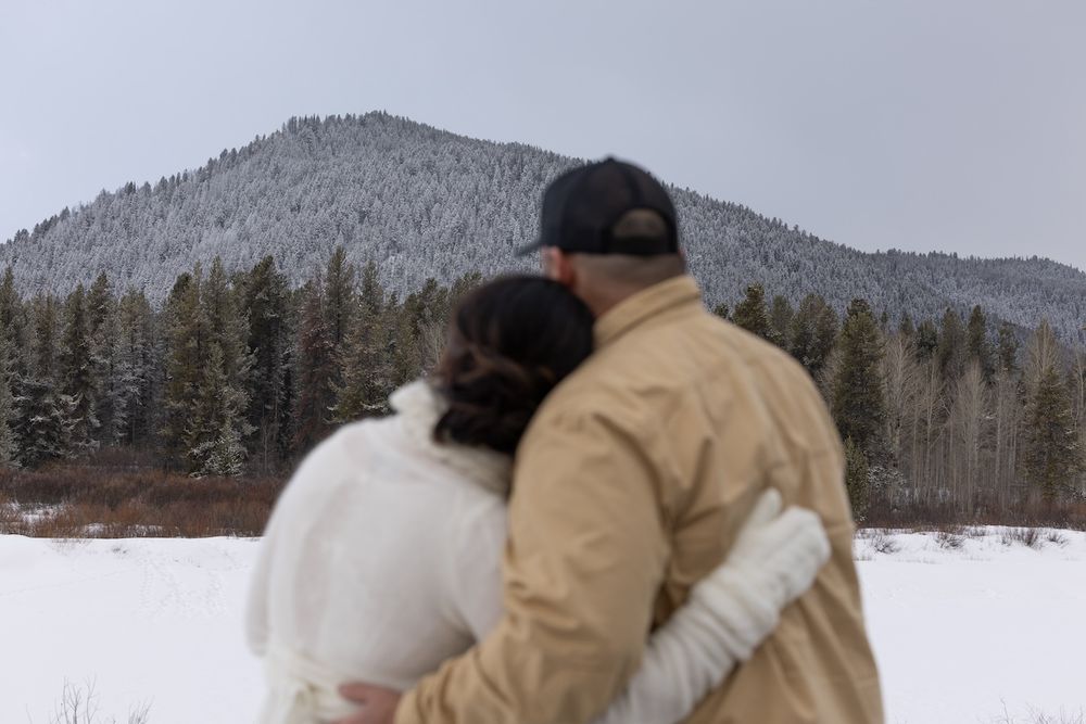A couple in winter coats embraces while looking at a snowy mountain landscape on a cloudy day.