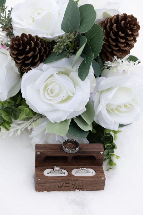 Wedding rings displayed in wooden box with white roses and pinecones in elegant floral arrangement above.