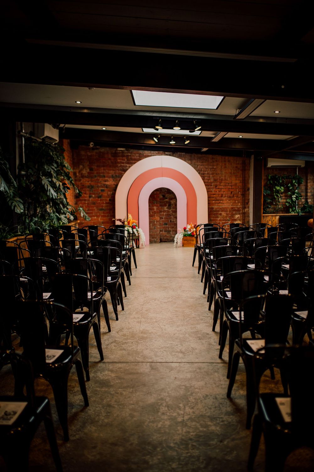 Dark industrial workspace with rows of black desks leading to an illuminated pink archway in a brick-walled room.
