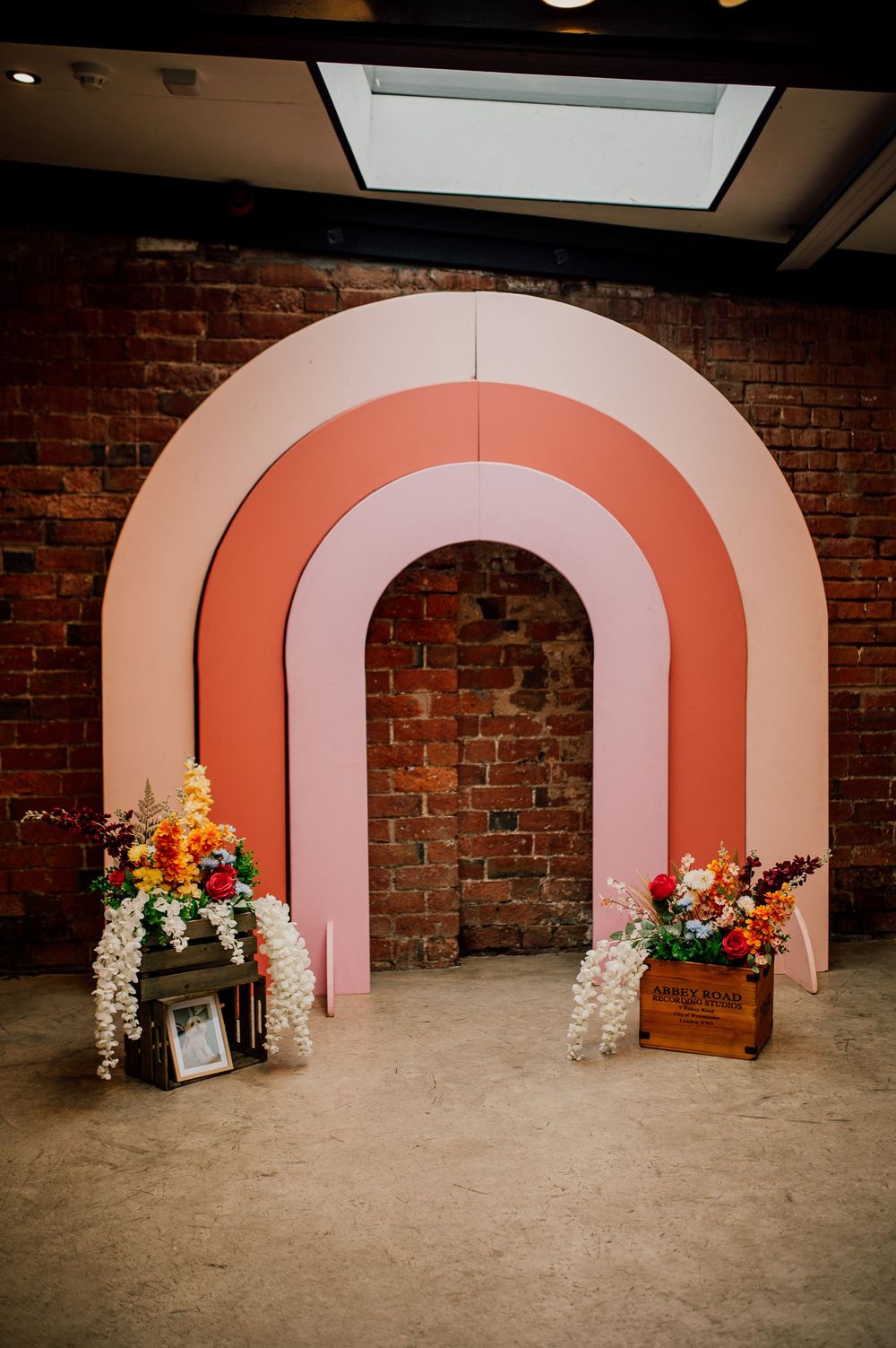 Modern pink and coral rainbow arch backdrop with floral arrangements in wooden boxes against exposed brick wall.