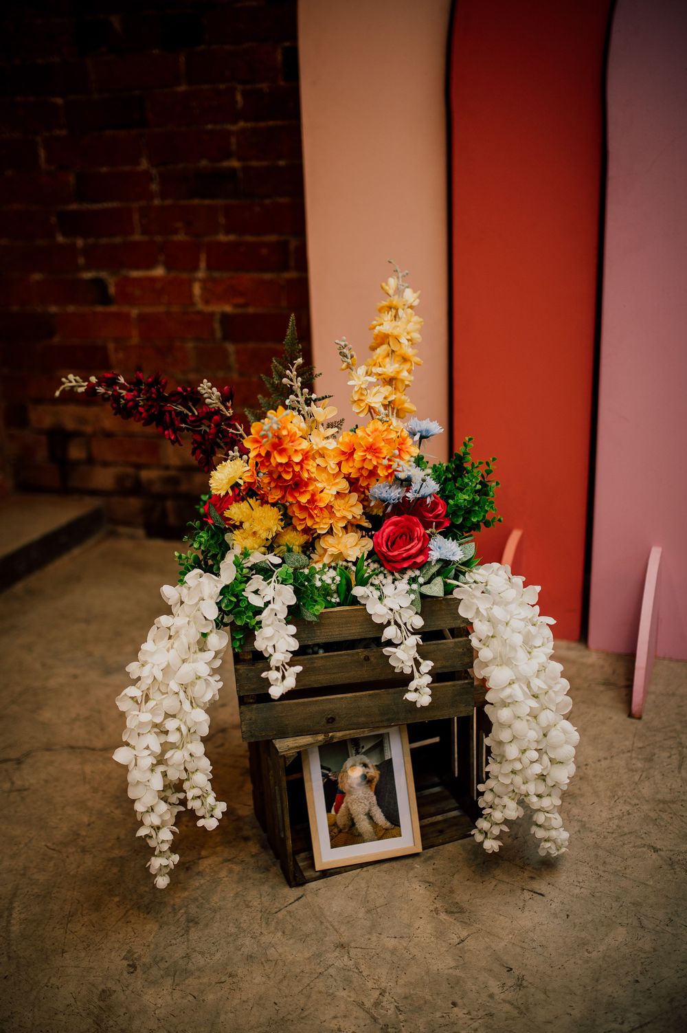 Rustic wooden crate decorated with cascading white orchids and vibrant orange and red flowers against a brick wall.