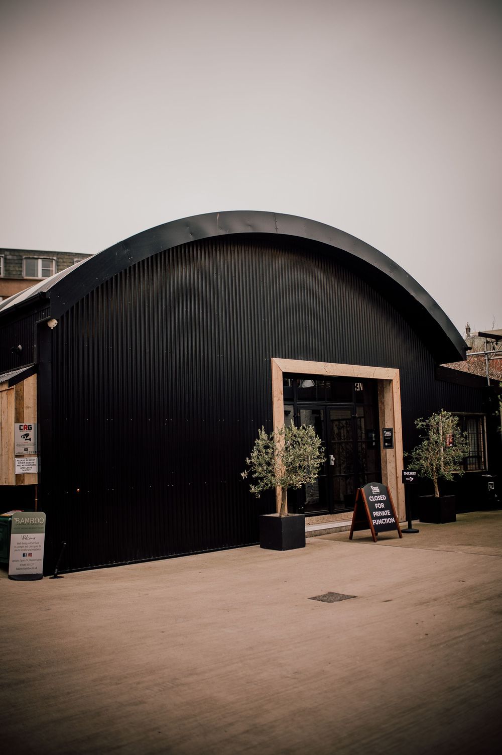 Modern black curved warehouse buildings with wooden accents line a concrete courtyard in an industrial setting.