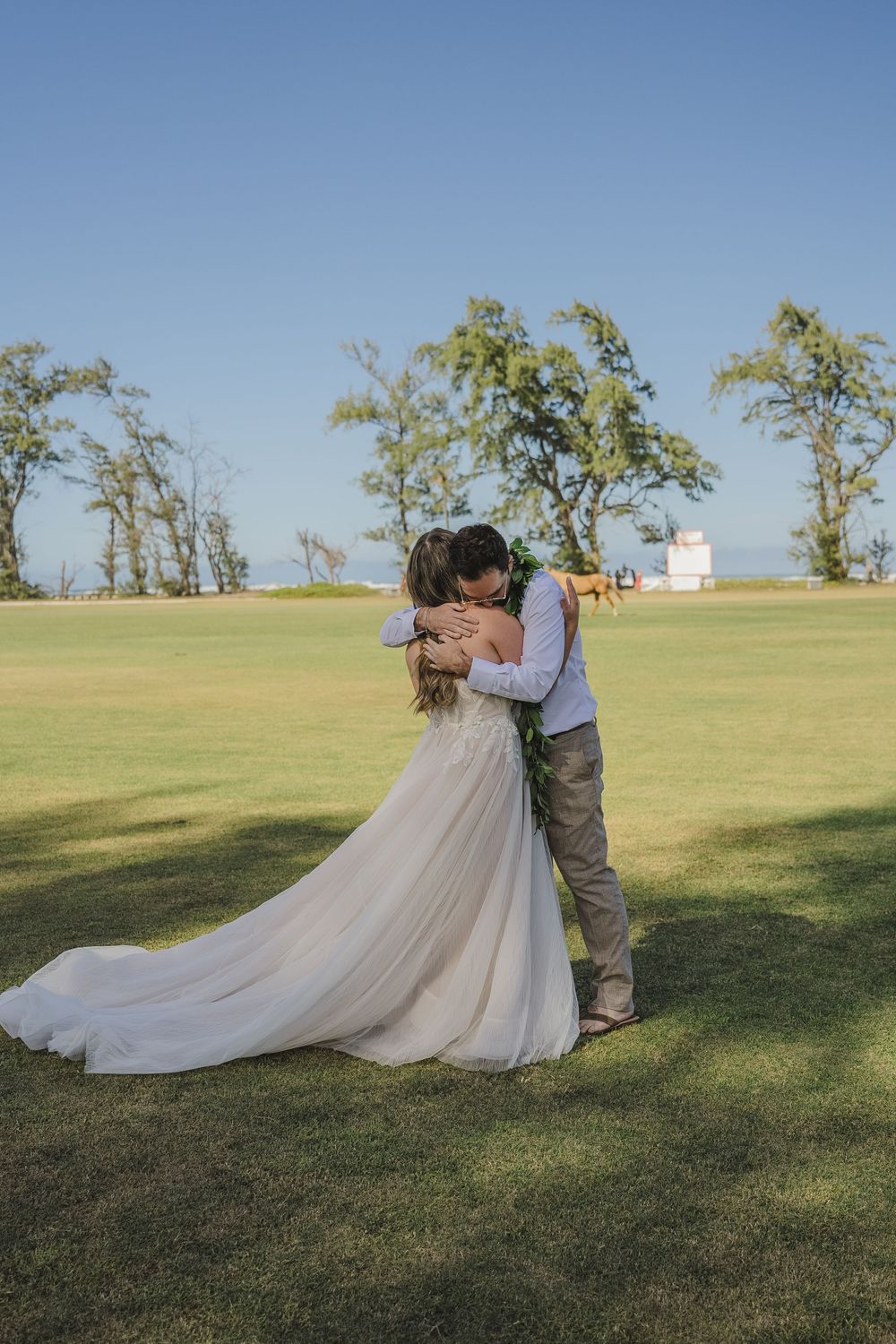A romantic wedding couple embraces on a hilltop lawn with flowing white dress and blue sky backdrop.