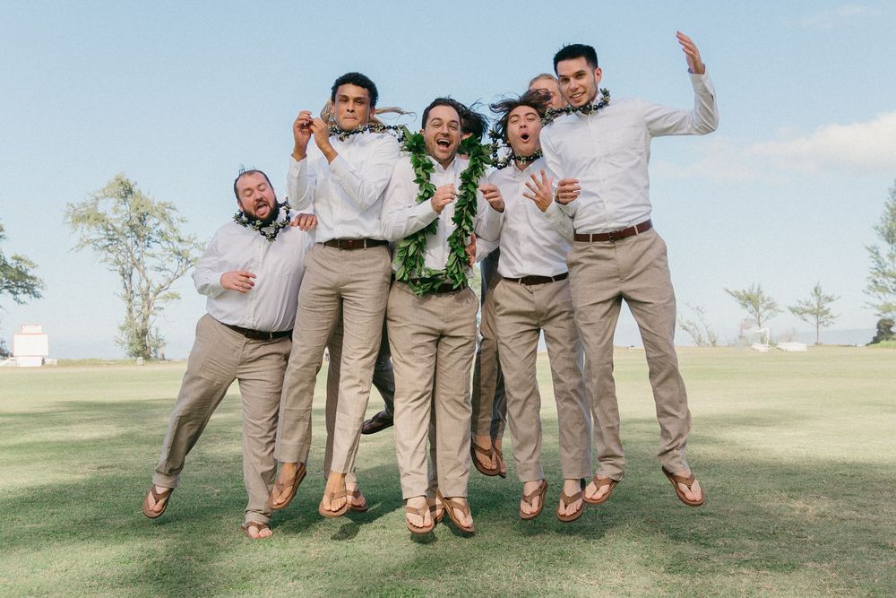 Groomsmen in tan suits and coral ties strike playful jumping poses during outdoor wedding photo session.