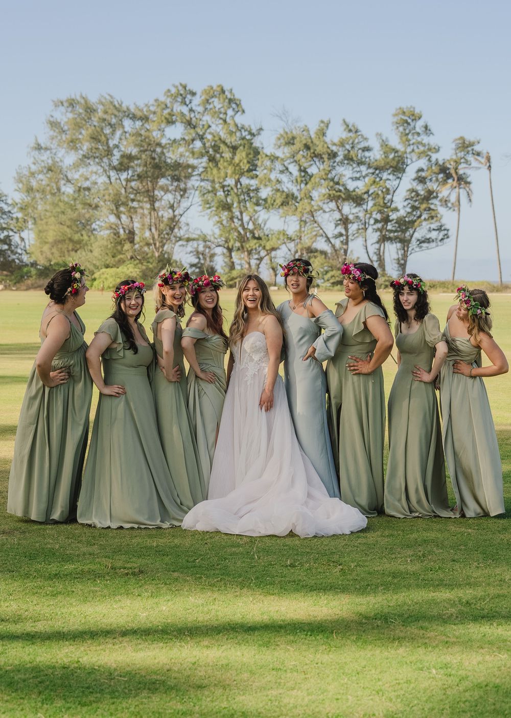 Wedding party in sage green dresses gathered on a sunny grassy hill for outdoor photos.