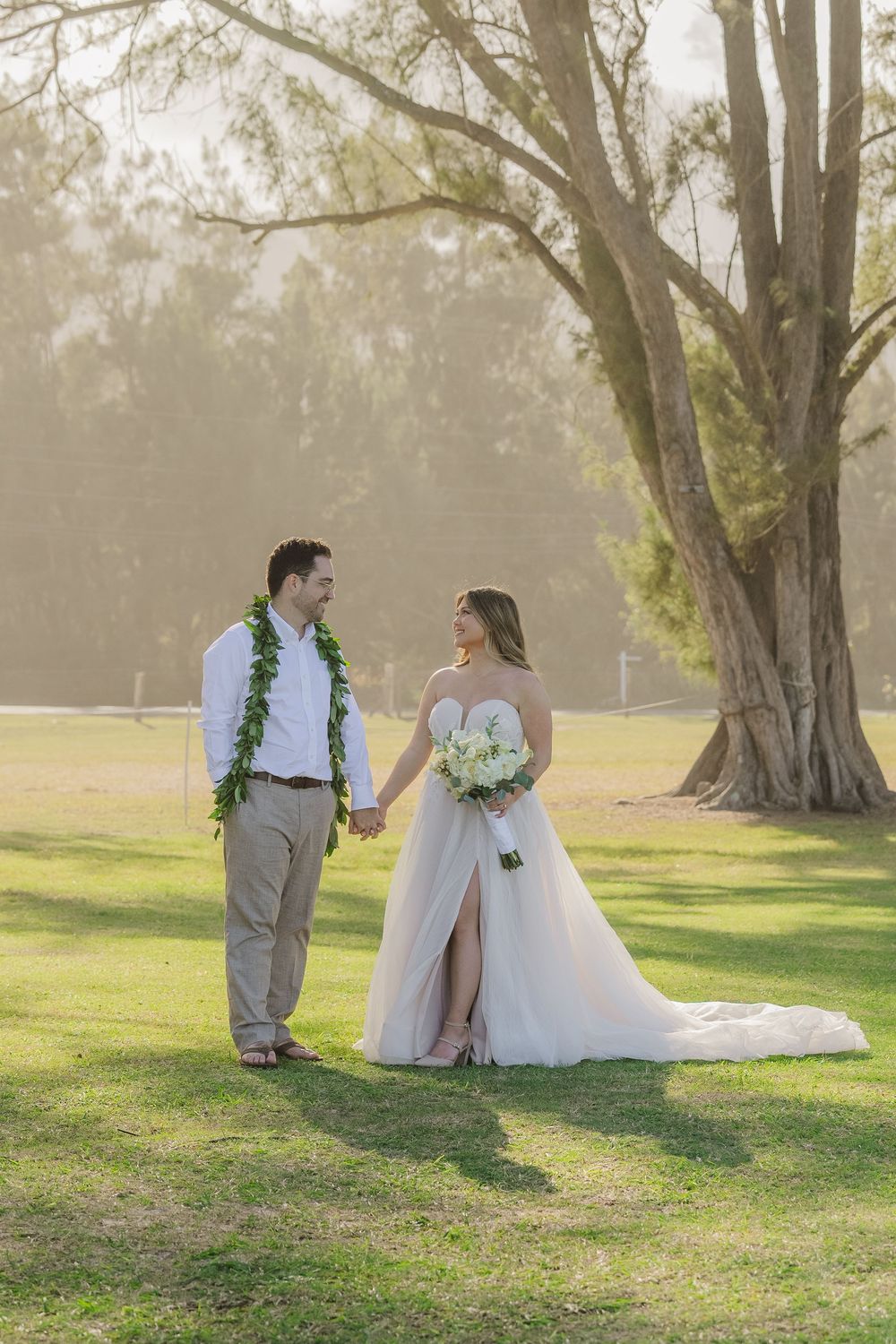 A couple in wedding attire stands together on a sunlit lawn with tall trees in the background.