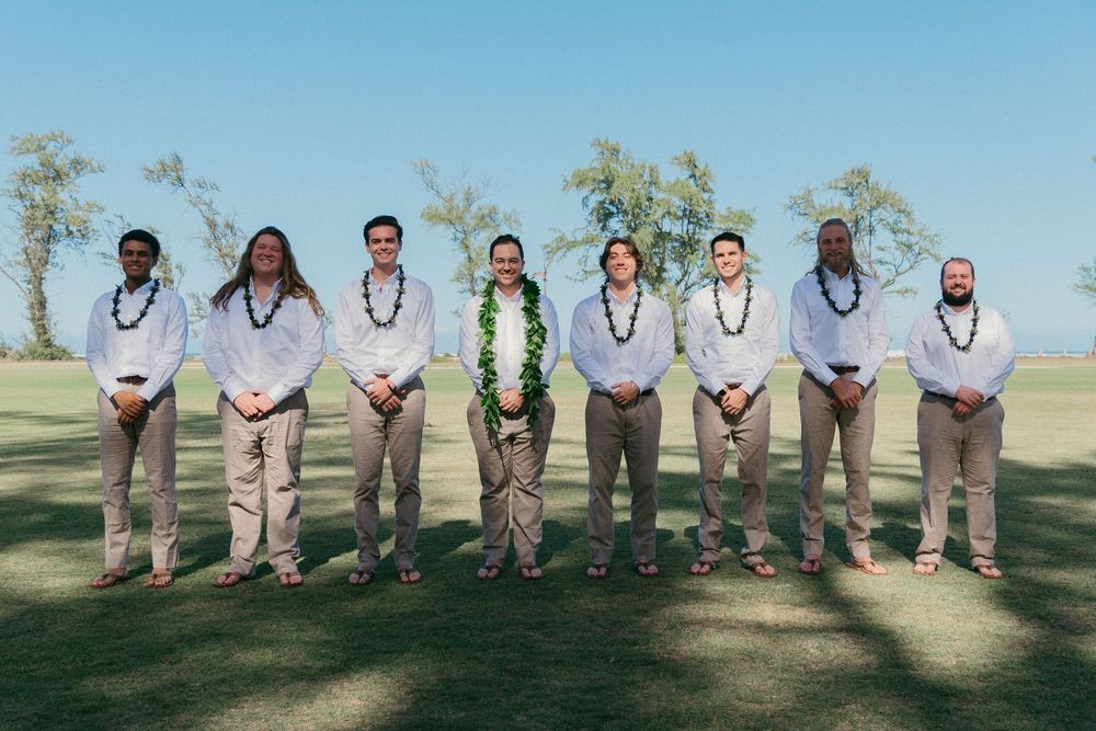 Wedding party in khaki suits and blue ties pose together on a grassy hill against a bright blue sky.