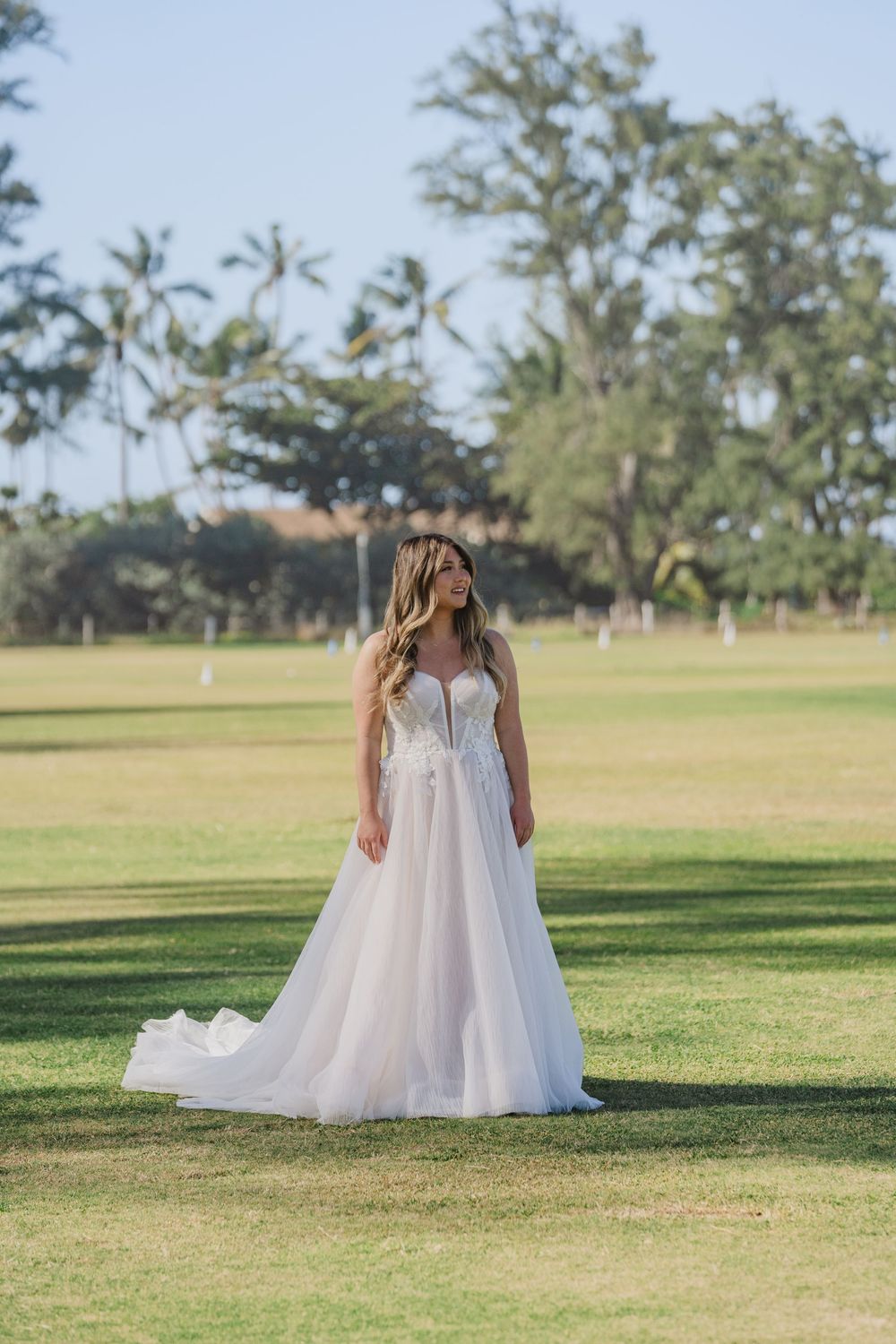 A bride in a flowing white gown poses on a grassy field with palm trees in the distance.