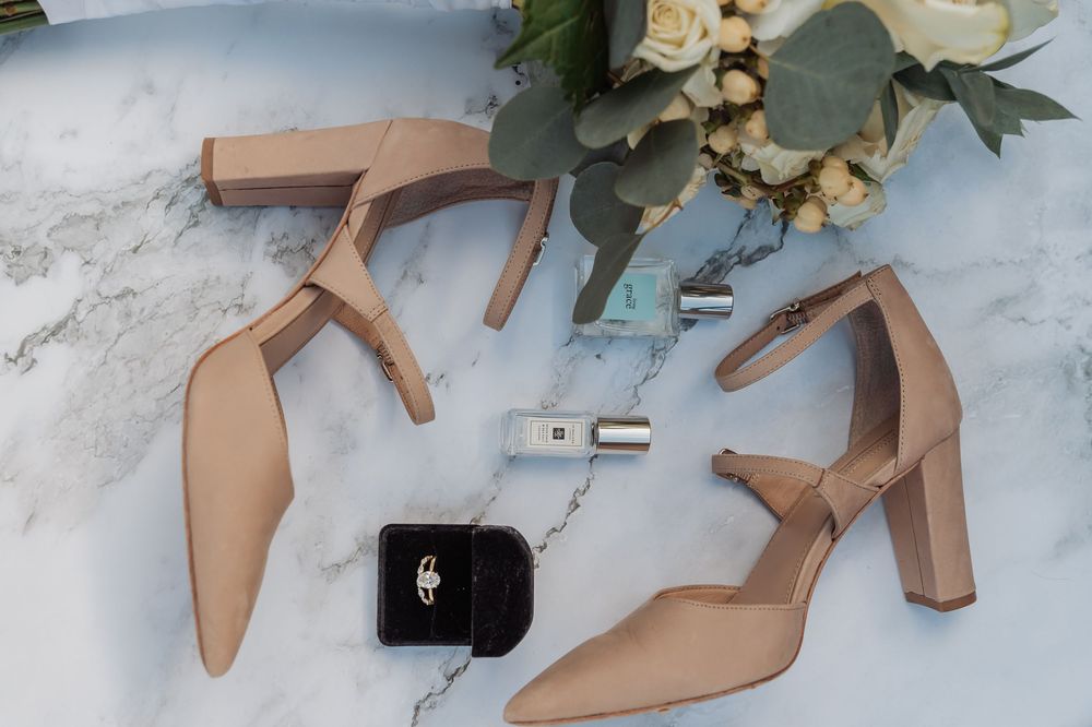 Nude leather ankle strap heels with pointed toe arranged with eucalyptus leaves and small black purse on marble surface.