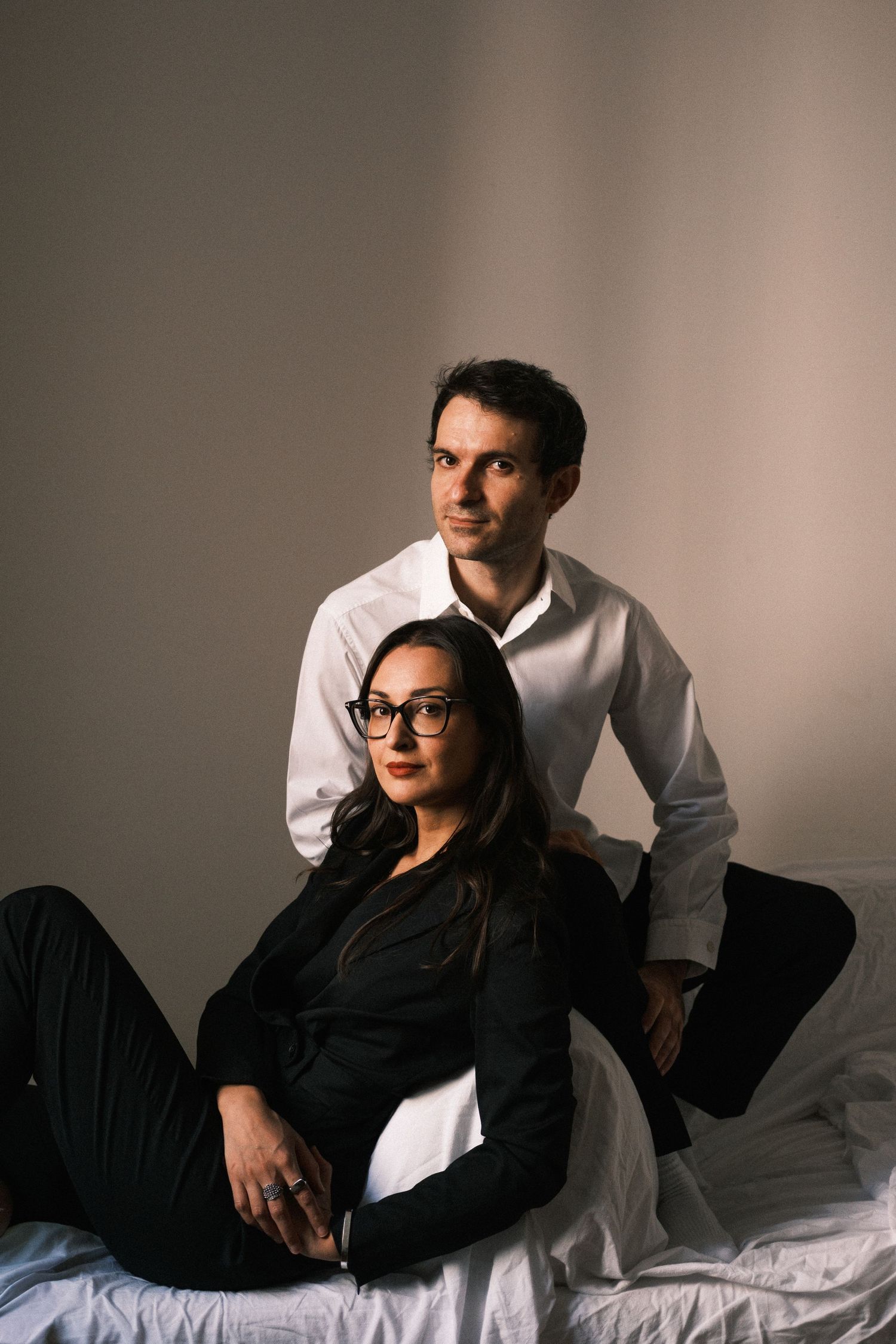 A couple poses together in a moody portrait, with one sitting on a bed while the other stands behind in white shirt.