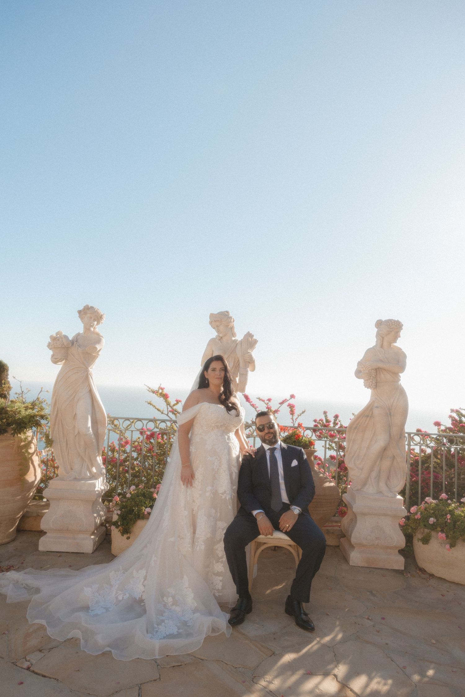 A bride and groom pose together on a Mediterranean terrace with classical statues and flowers at sunset.