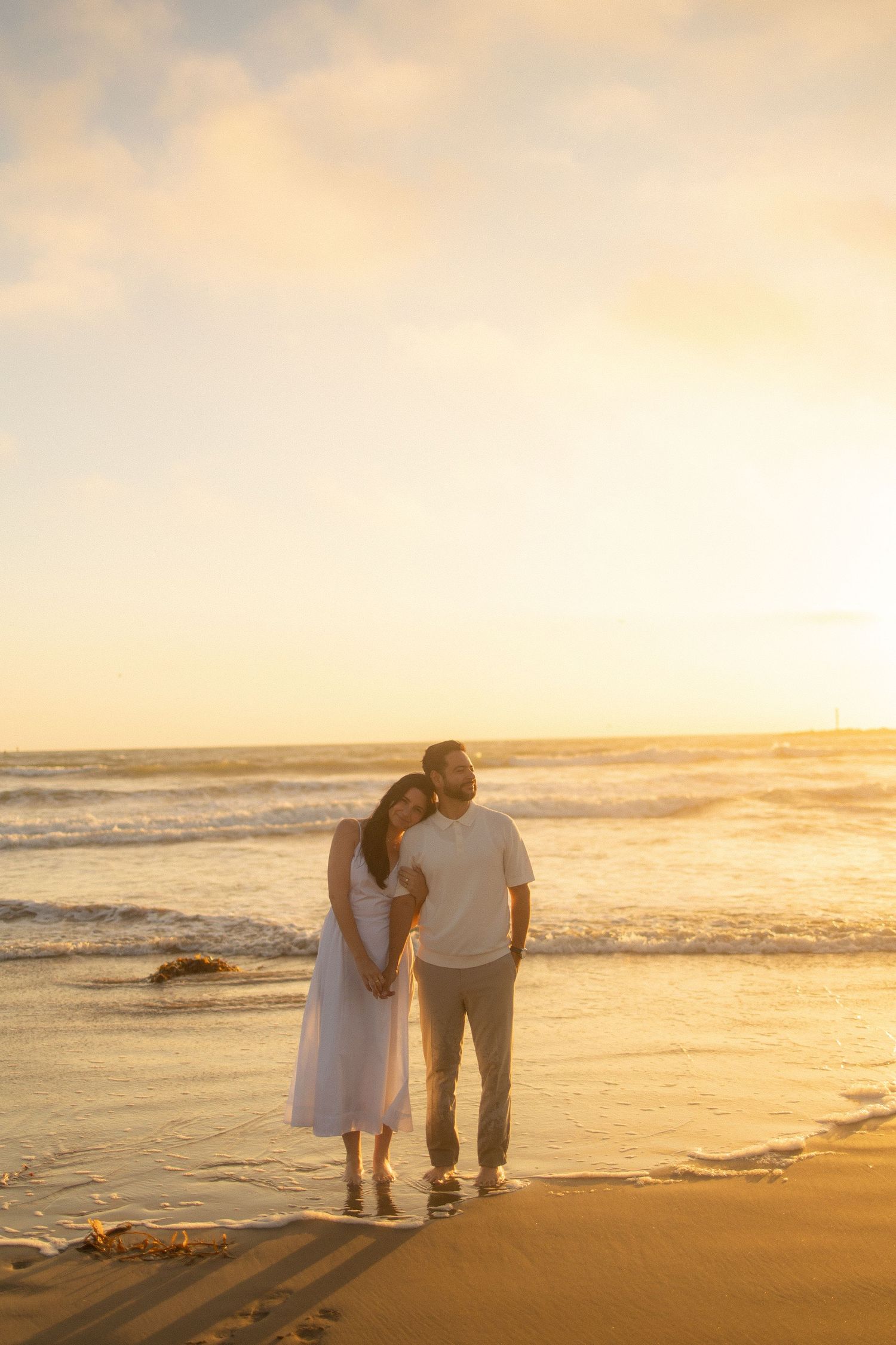 A couple stands together on a sandy beach at sunset as gentle waves roll in along the shoreline.