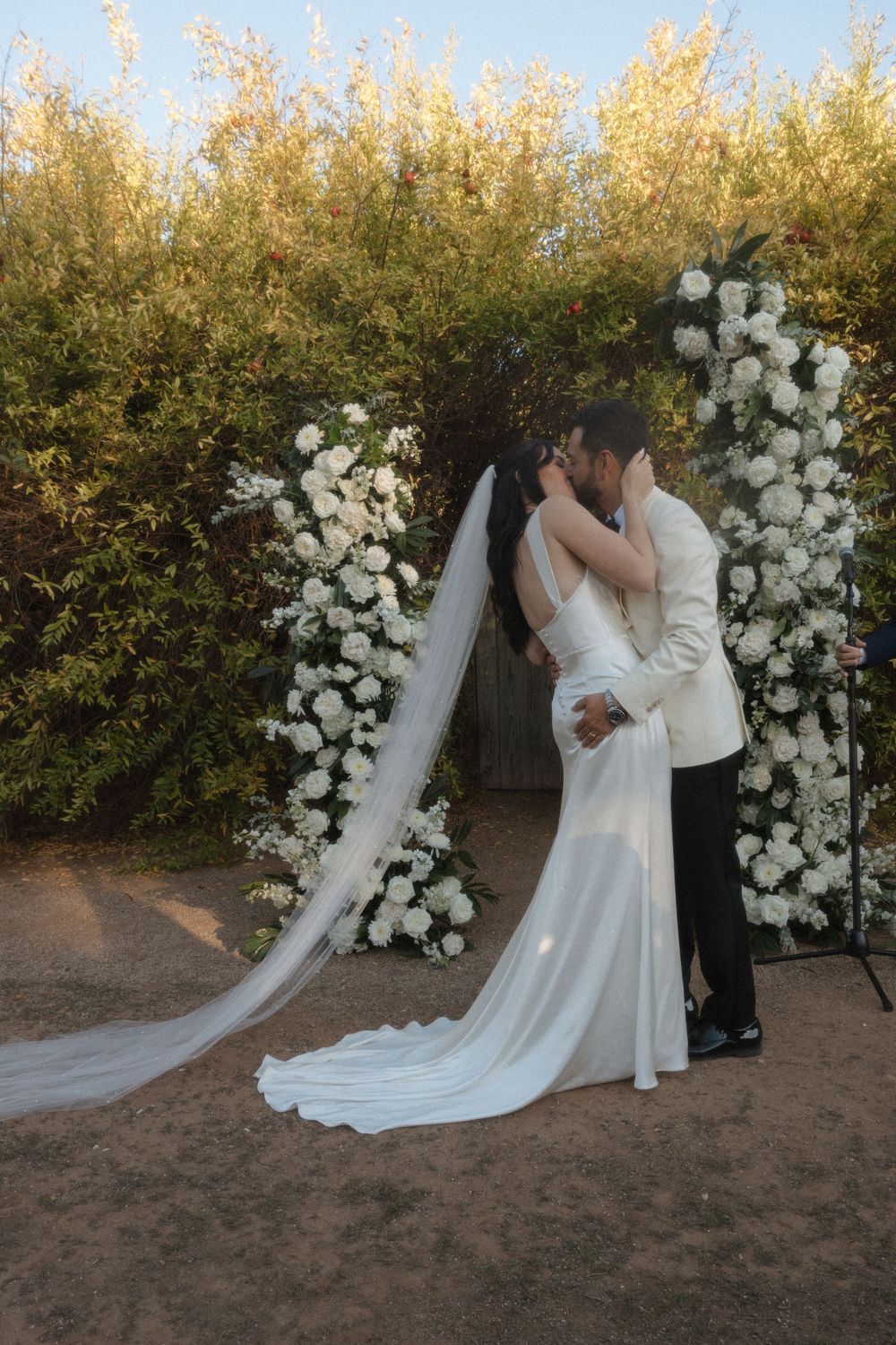 Romantic wedding couple shares a kiss under a floral arch decorated with white roses at sunset.