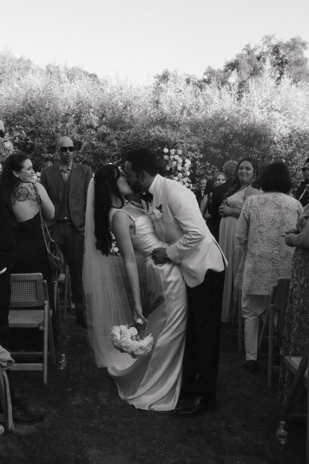 A romantic black and white wedding moment captured outdoors with guests looking on as couple shares an intimate kiss.
