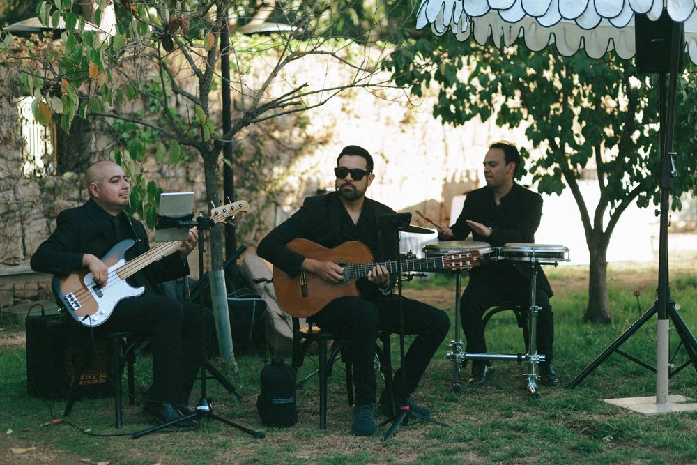 Three musicians perform outdoors with acoustic guitar, bass guitar and percussion under a shaded garden setting.