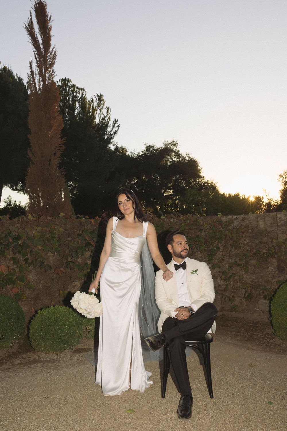 A couple poses outdoors at sunset, with the bride in a white gown holding flowers and groom seated in formal attire.