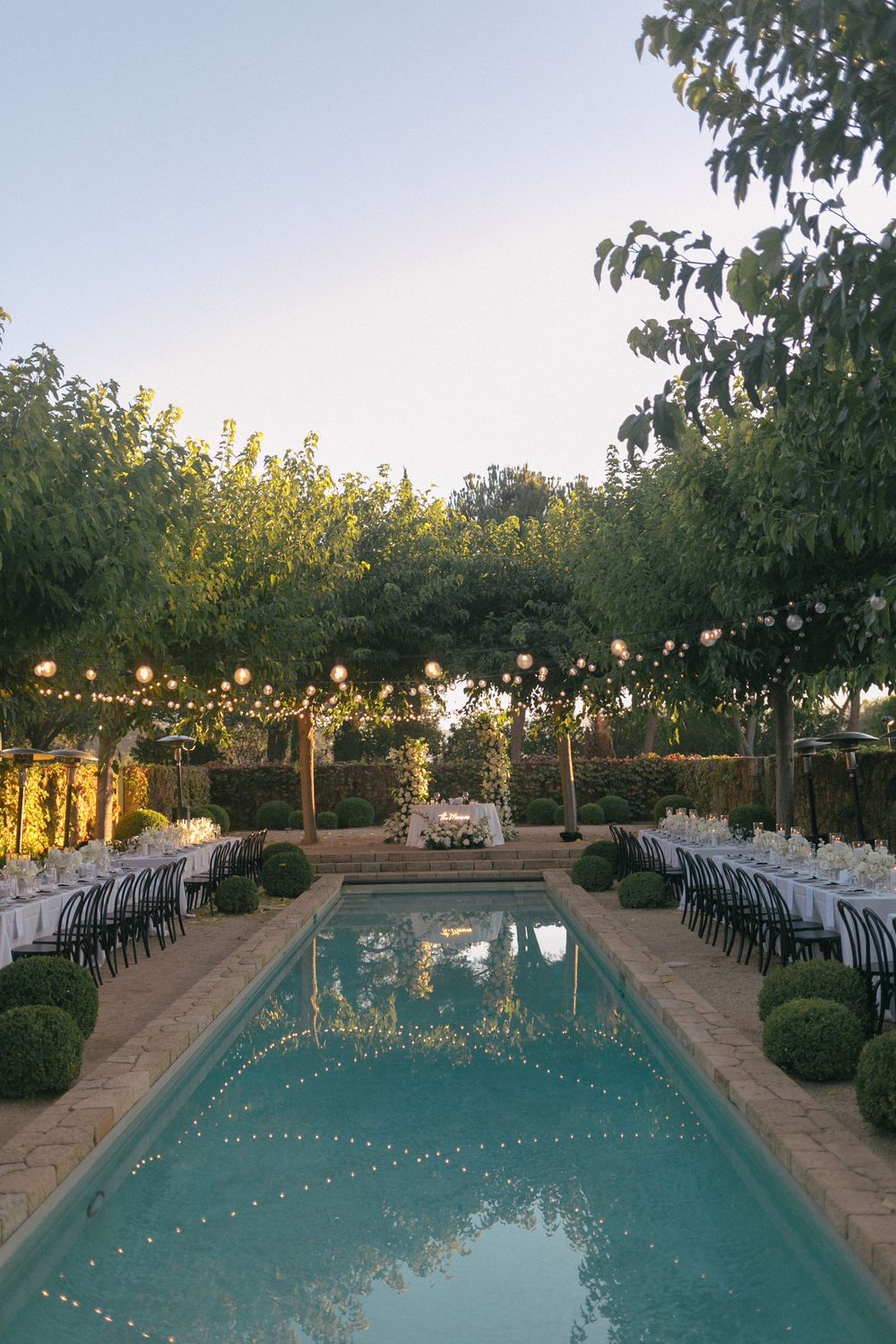 Long tables set for an outdoor dinner party beside a swimming pool with string lights hanging overhead at sunset.