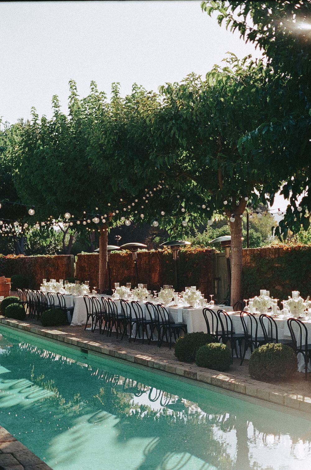 Long outdoor dining tables set for an elegant wedding reception beside a turquoise swimming pool with string lights overhead.