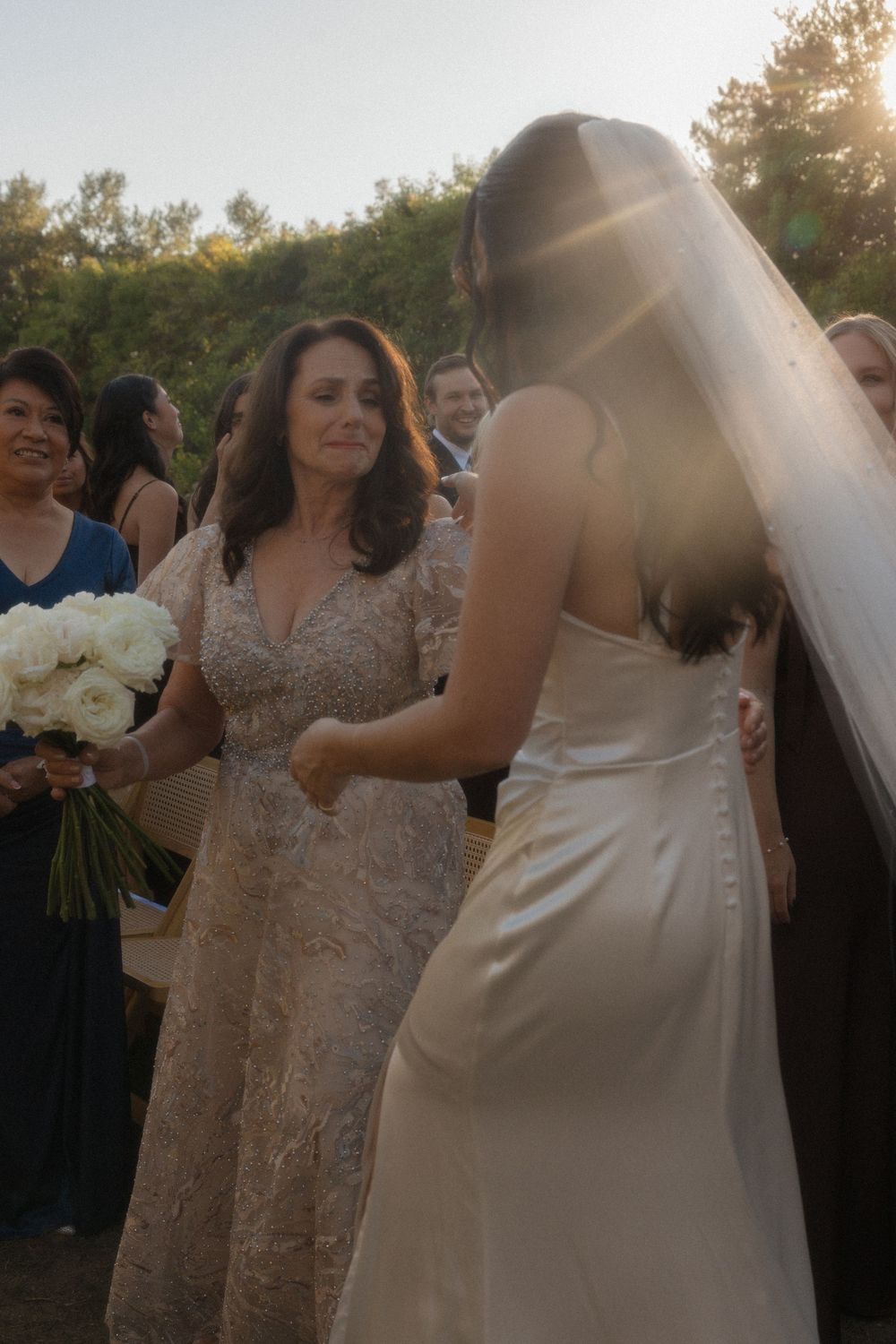 A joyful outdoor wedding celebration with people in formal attire and white floral bouquets at sunset.