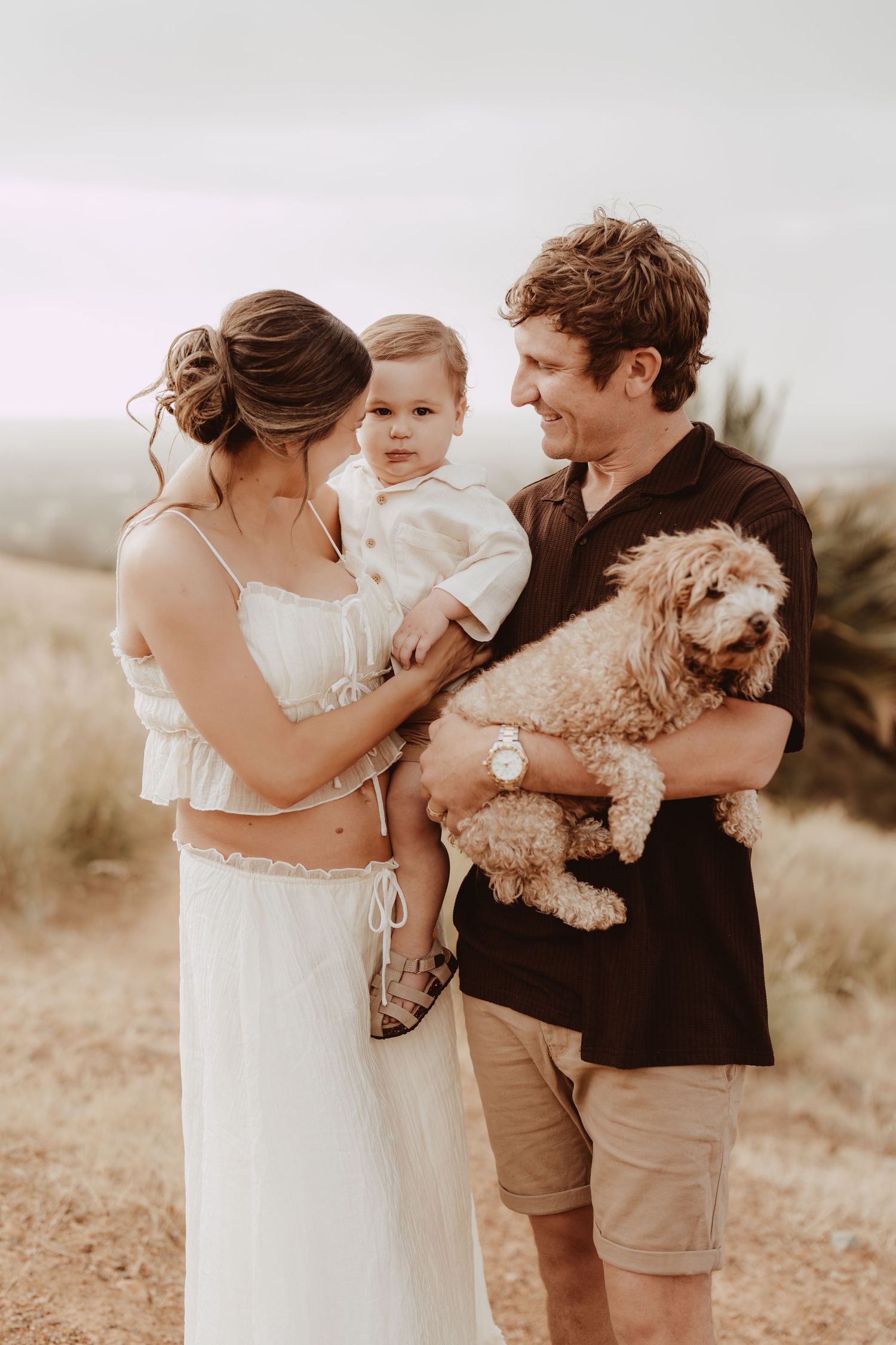 A family in casual attire poses together outdoors with their small dog in a warm-toned natural setting.