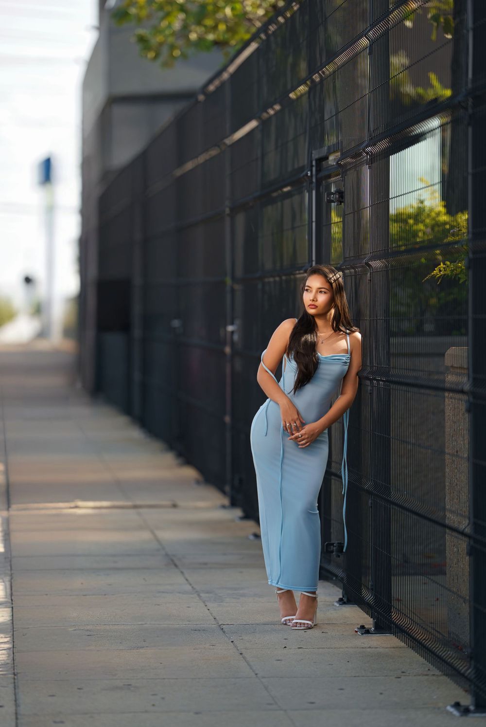 A woman in a light blue fitted maxi dress poses against a modern glass building exterior on a sunny day.