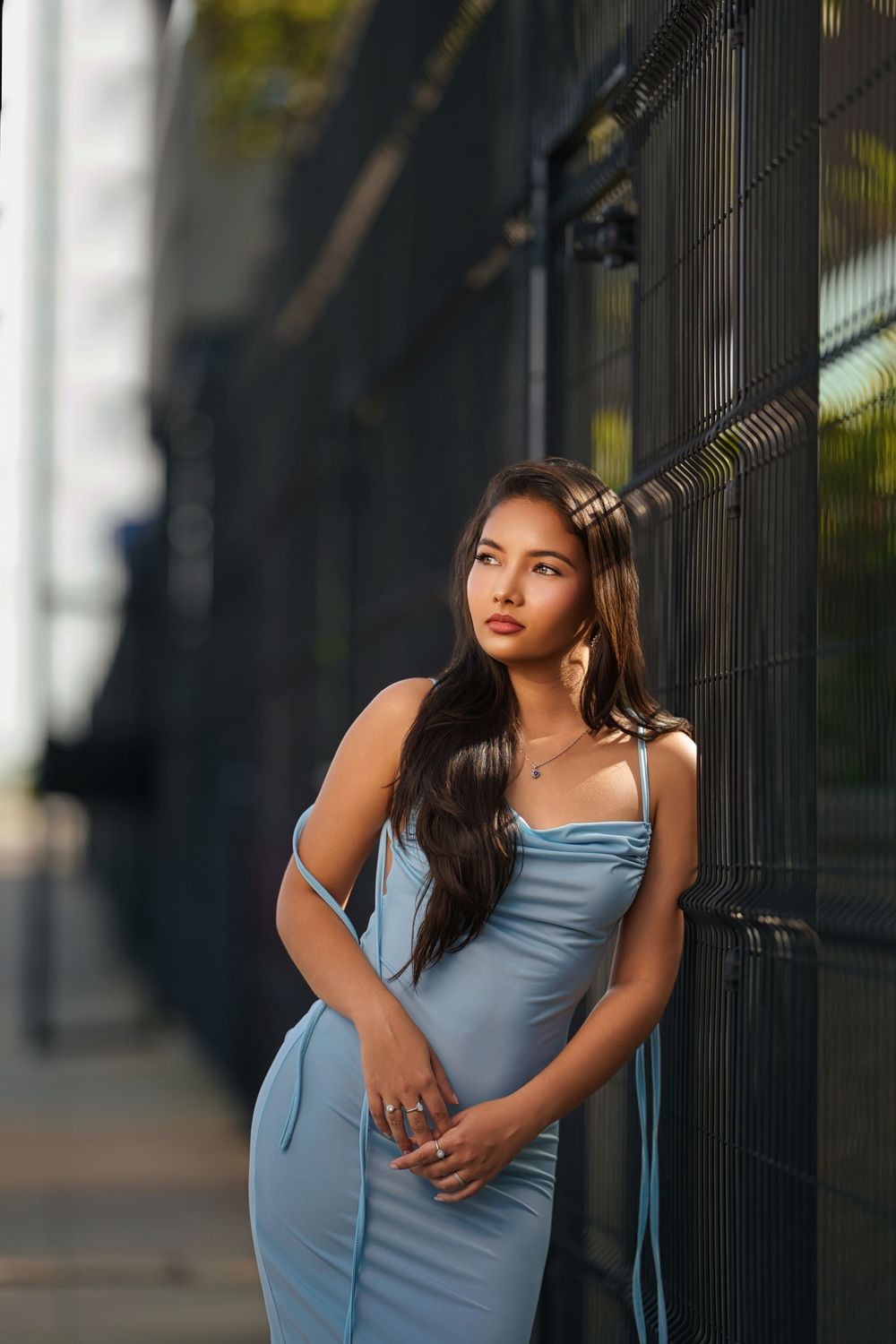 A person in a light blue dress poses elegantly against a black metal fence in an urban setting.