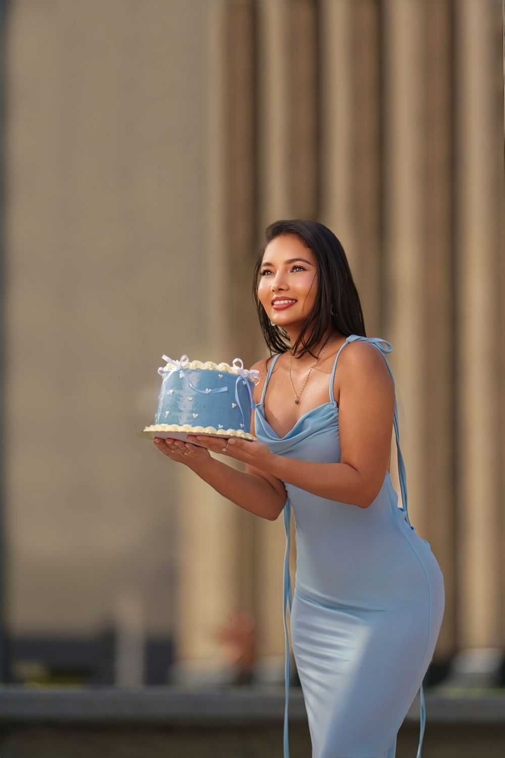A woman in a blue dress holds a decorated cake while smiling during a celebration photoshoot.