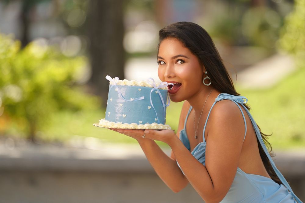 Someone in a blue top playfully bites into a decorated light blue cake outdoors on a sunny day.